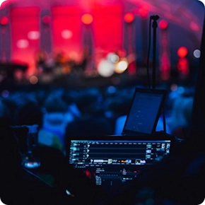 Een concertscène met een wazig publiek, felrode verlichting op het podium en een opvallende geluidstechnicus op de voorgrond. De sfeer is energiek en levendig.