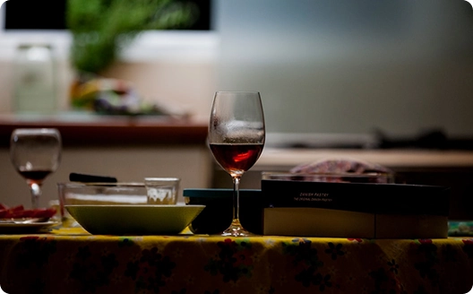 Op een slecht verlichte tafel met een bloemetjeskleed staan twee wijnglazen, waarvan er één gedeeltelijk gevuld is met rode wijn, een kom, borden en een wazige keuken op de achtergrond.