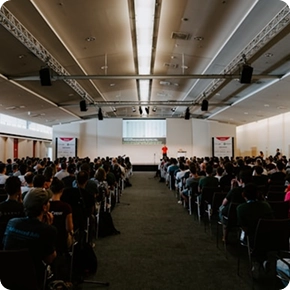 Twee basketbalspelers in volle actie op het veld: de ene, in het wit, speelt de bal door, terwijl de andere, in het blauw, verdedigt. De zaal zit bomvol, wat voor een elektrische sfeer zorgt.