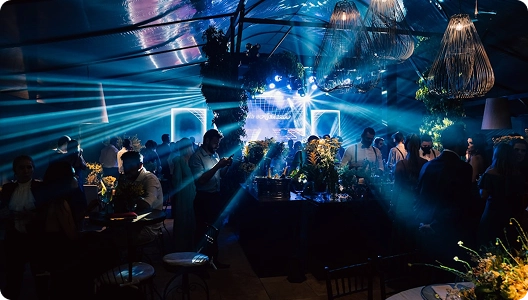 Een levendig feest met felblauwe verlichting en decoratieve hanglampen. De gasten staan gezellig bij elkaar rond tafels met kleurrijke bloemstukken, wat zorgt voor een energieke en feestelijke sfeer.