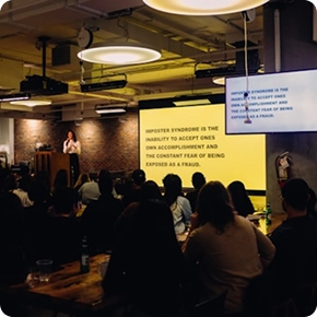 Een spreker houdt een presentatie voor een zittend publiek in een schemerige ruimte. Op een groot scherm wordt tekst over het impostorsyndroom getoond. De sfeer is geconcentreerd en aandachtig.