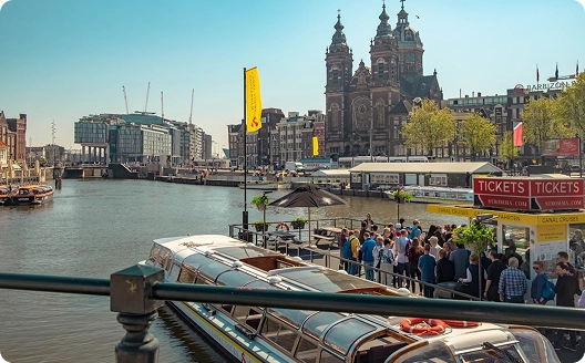 Een zonnige dag in Amsterdam, met een gracht vol boten, de Sint-Nicolaaskerk op de achtergrond, een menigte die kaartjes koopt en historische gebouwen in de buurt.