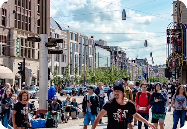 Een drukke straat in de stad met een bonte mengeling van voorbijgangers onder een blauwe hemel. Mensen lopen rond en gaan met elkaar in gesprek, omringd door historische en moderne gebouwen. Er hangt een levendige sfeer.