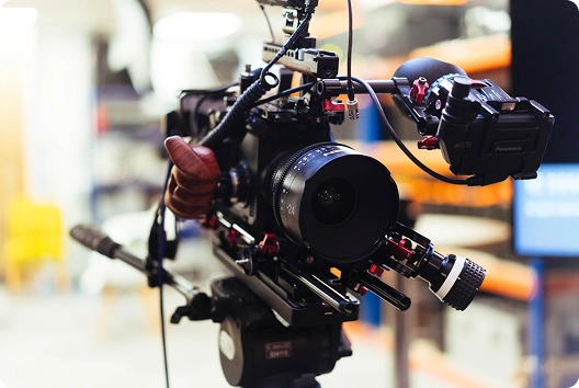 Close-up van een professionele videocamera-opstelling met diverse accessoires en een grote lens scherp in beeld. De achtergrond is wazig, wat doet vermoeden dat de opname binnenshuis is gemaakt.