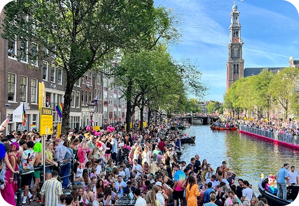 Een drukke straat in de stad met een bonte mengeling van voorbijgangers onder een blauwe hemel. Mensen lopen rond en gaan met elkaar in gesprek, omringd door historische en moderne gebouwen. Er hangt een levendige sfeer.