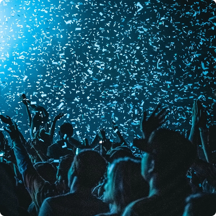 Een bruisend concert met silhouetten van een enthousiast publiek dat de handen in de lucht steekt onder een regen van confetti, verlicht door blauwe podiumlampen. Een energieke en feestelijke sfeer.